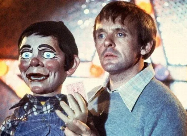 Still from Richard Attenborough, Magic, 1978. A man stares forlorn while holding a ventriloquist dummy. In the background are bright and colorful lights in the shape of a star.