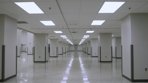 An empty office building interior with white tile floors and white pillars, shrouded in clinical LED lighting.